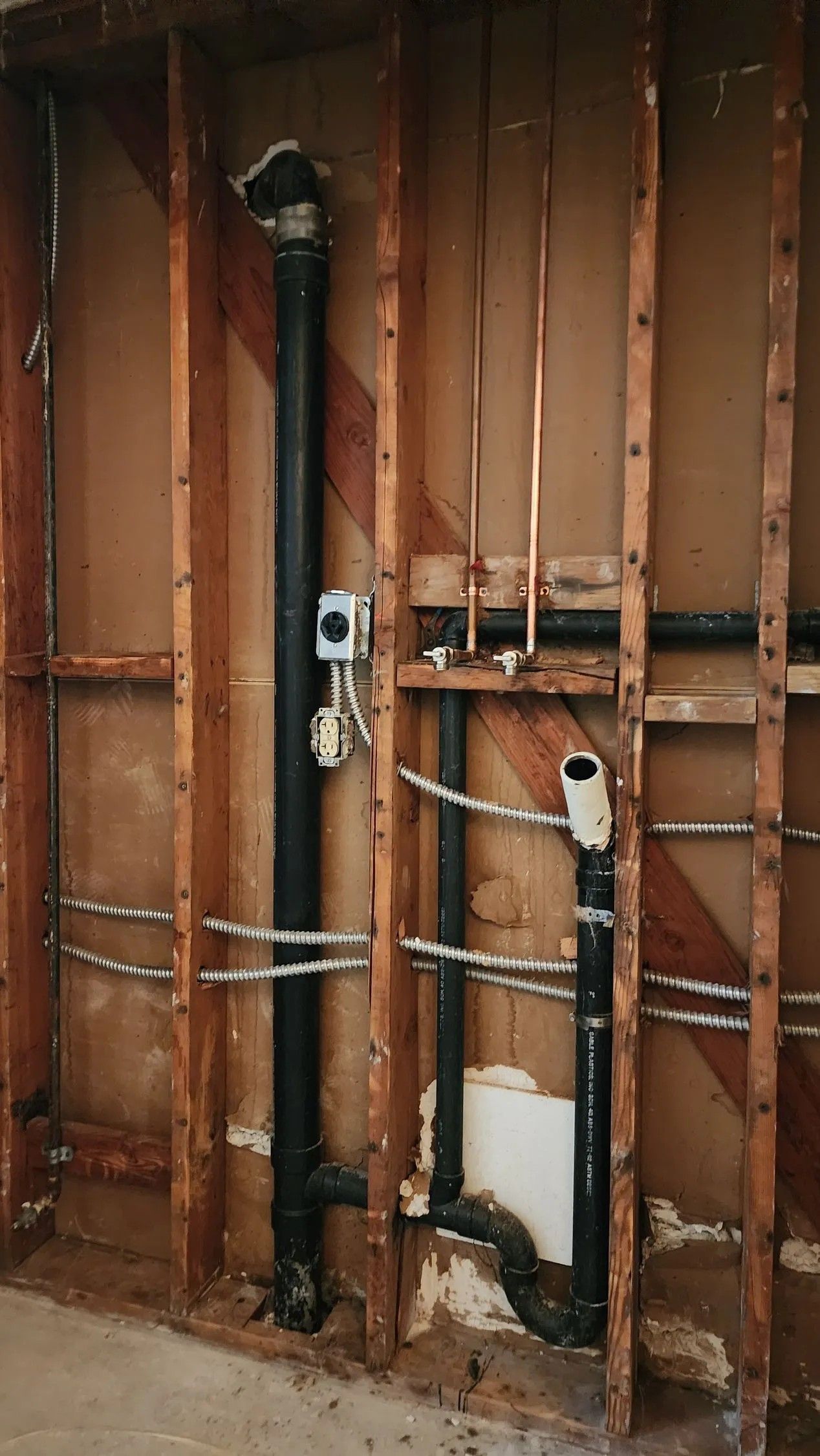 Plumbing pipes exposed in a wall. Black drain pipes, copper water lines, and wooden studs are visible.