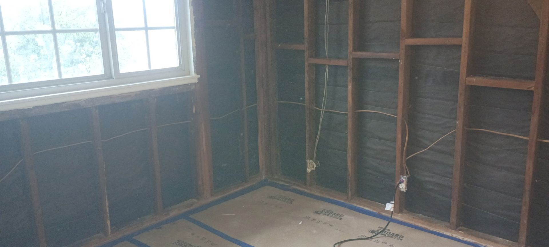 Interior of a room under construction with exposed wooden framing and wiring; a window is visible.