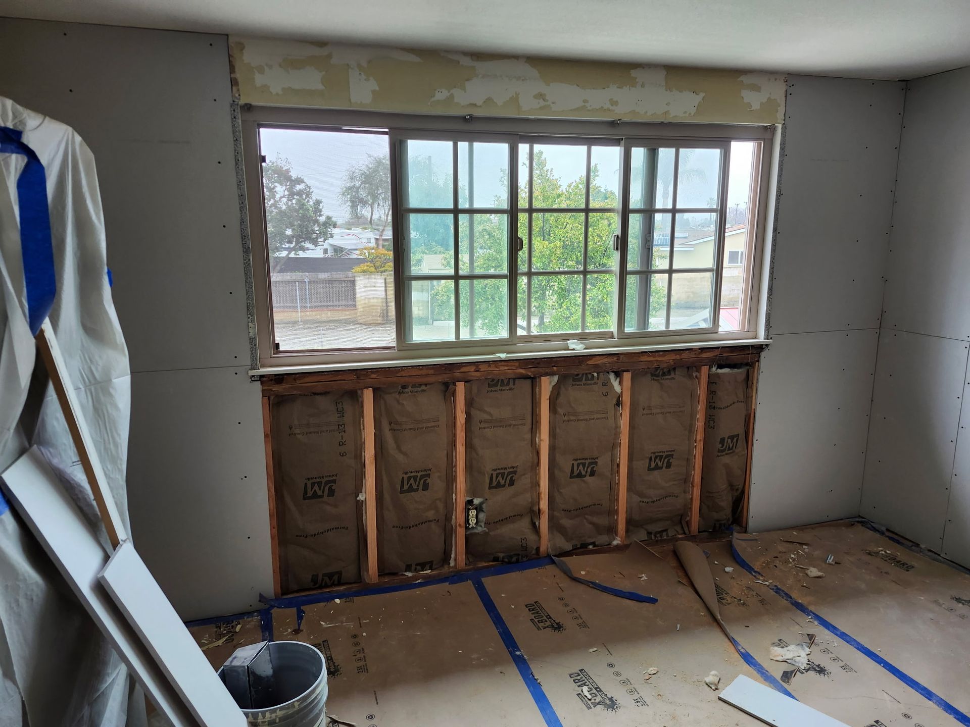 Room undergoing renovation with exposed wall studs, insulation, and a window overlooking a yard.