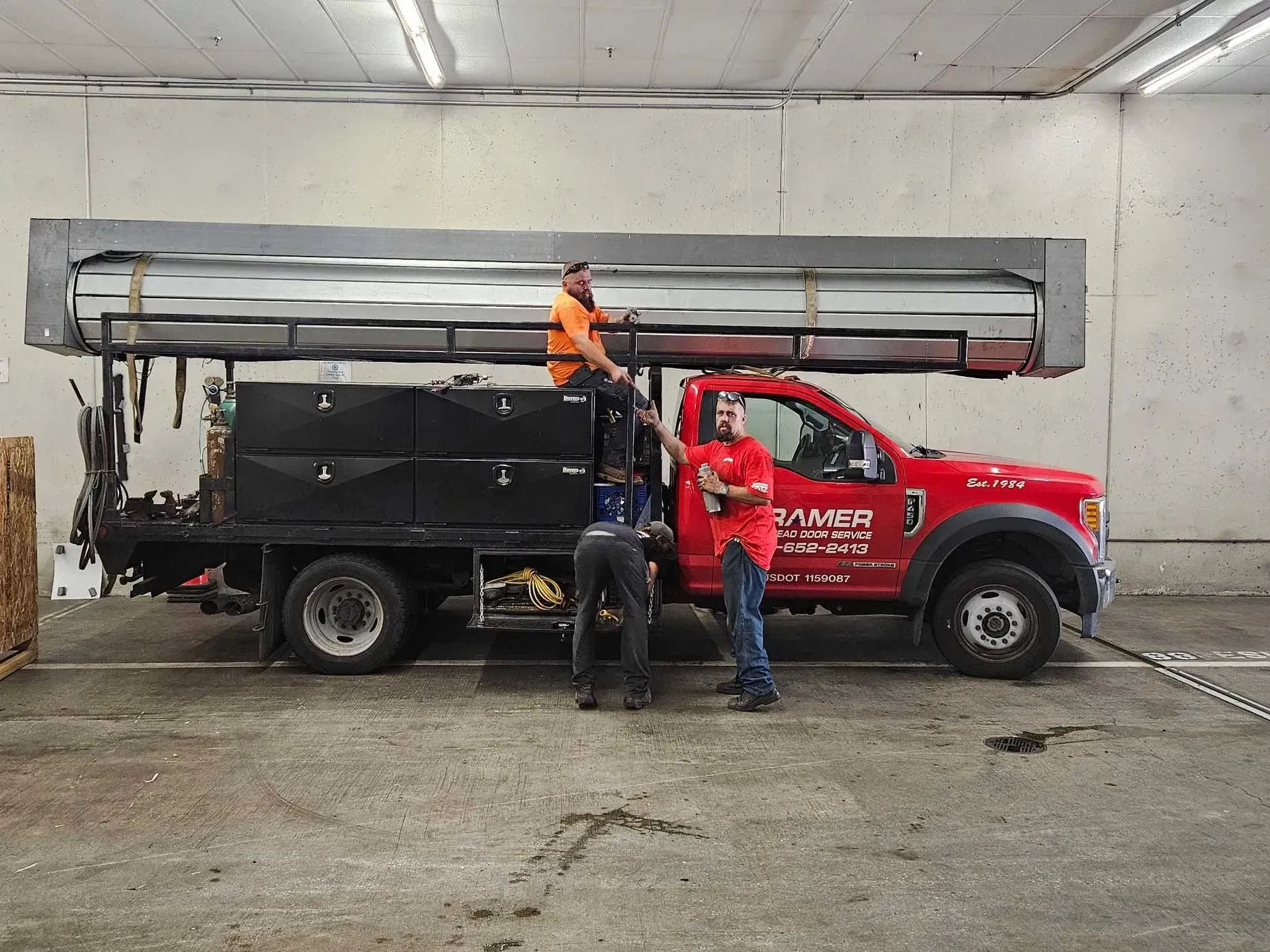 Three workers load a large metal structure onto a flatbed truck inside a warehouse.