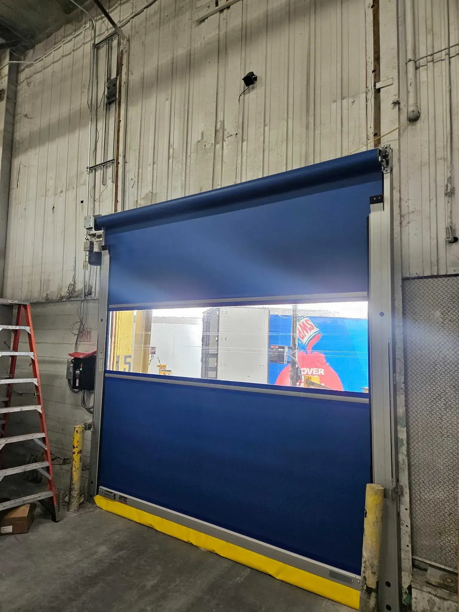 Blue industrial roll-up door partially open, revealing a warehouse interior; ladder on the left.