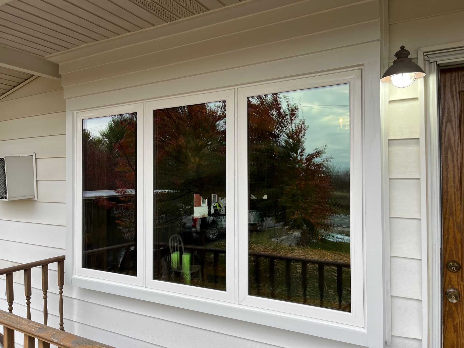 White-framed windows on a beige house exterior with porch railing and a lit wall lantern.