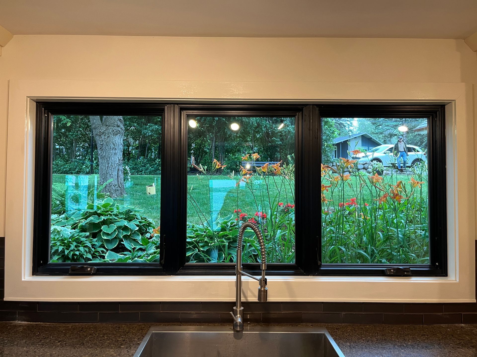 Kitchen window overlooking a green garden with flowers, framed in black trim above a sink faucet