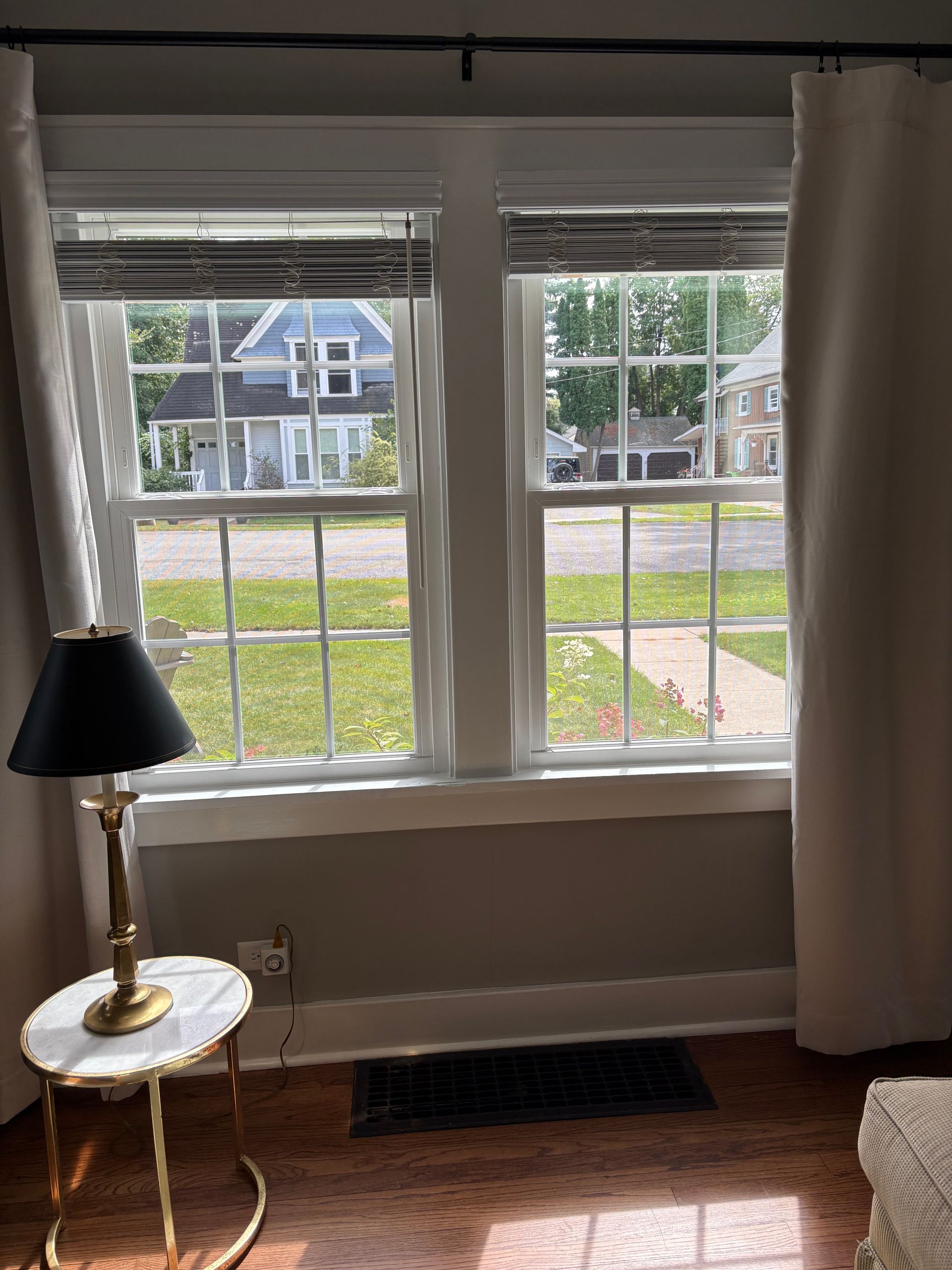 Living room window view with white curtains, lamp on side table, and a house across the street.