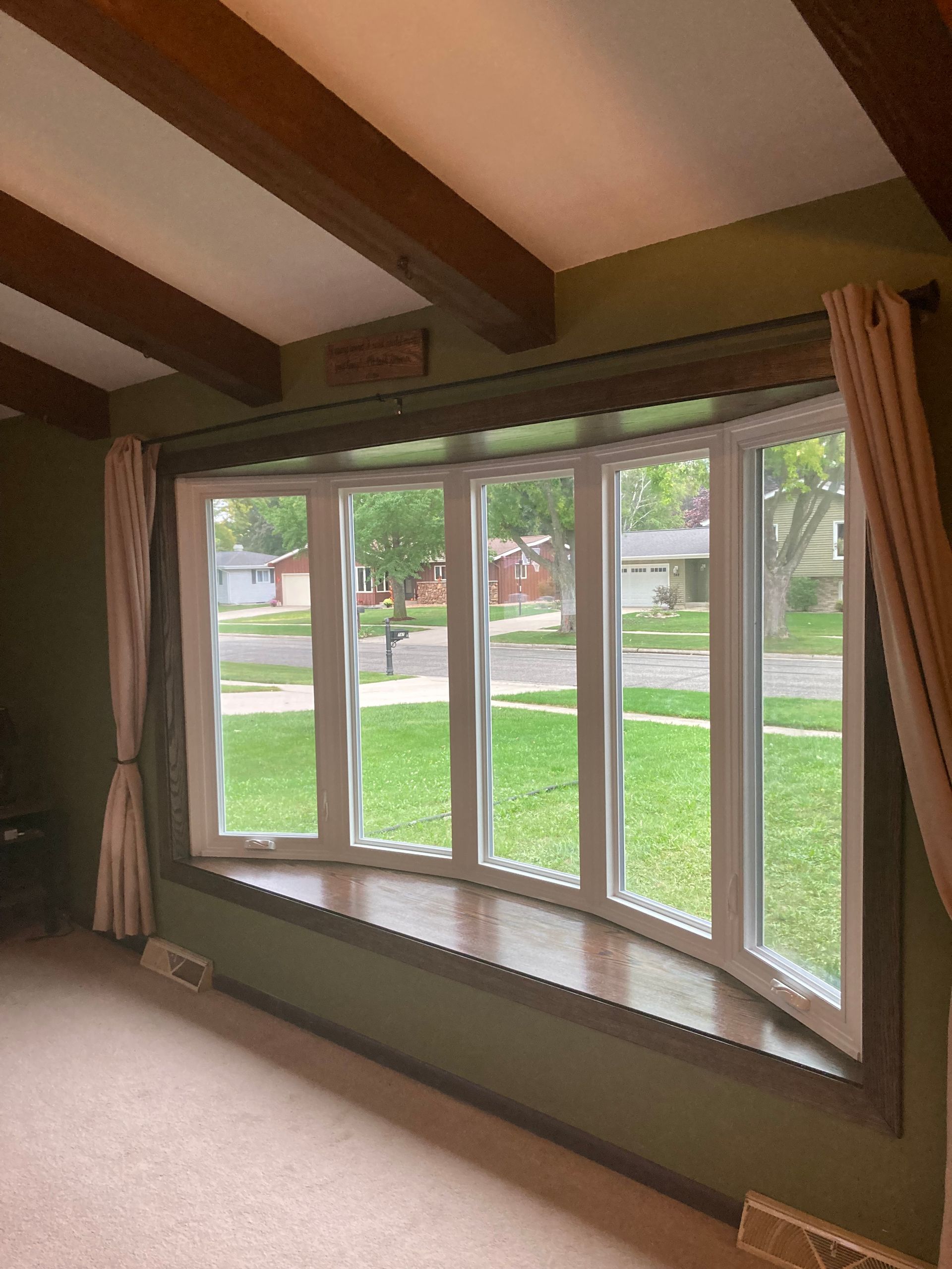 Living room bay window with beige curtains, wood beams, and a view of a suburban street and lawn