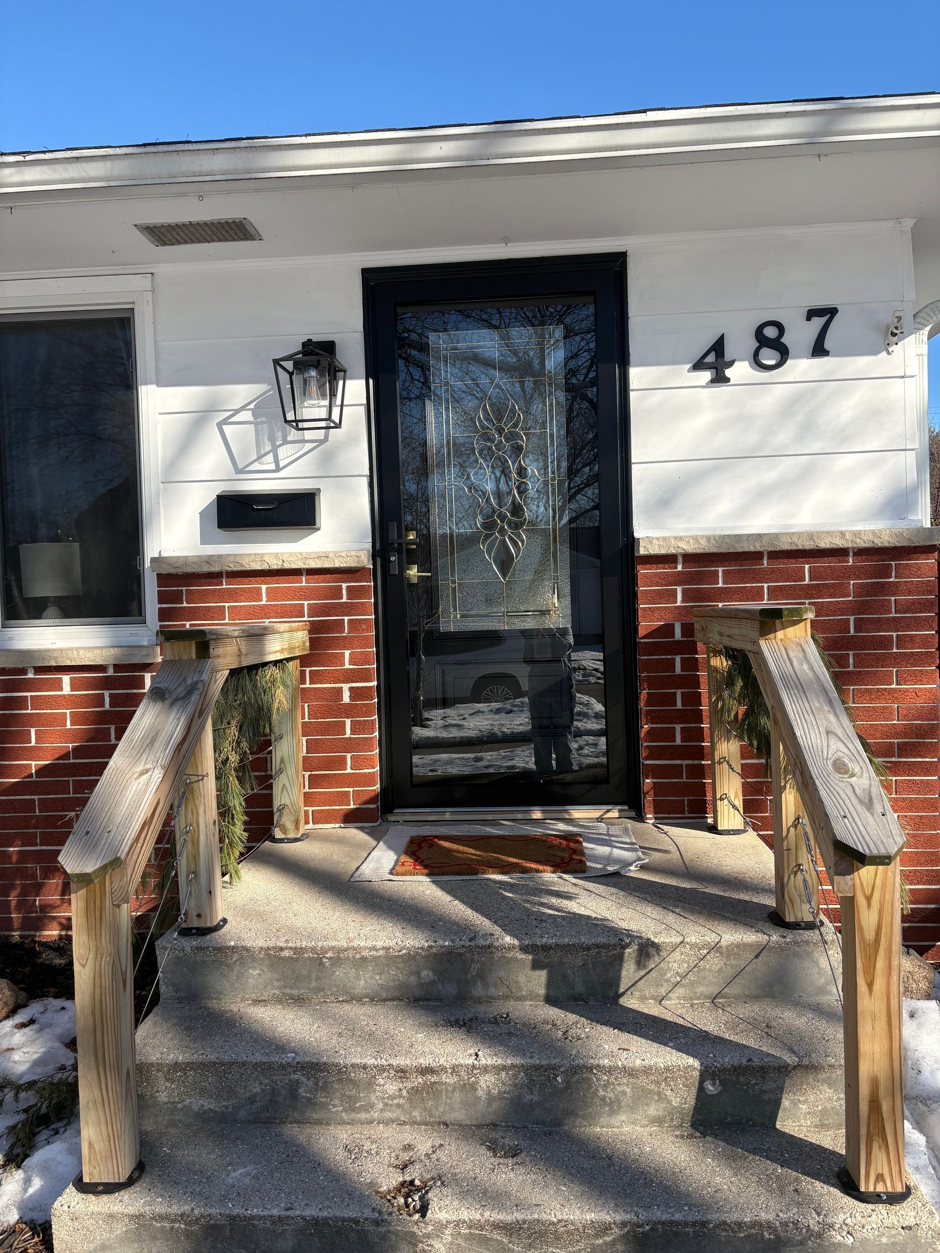 House entrance with black glass storm door, brick lower facade, and wooden accessibility ramp with handrails.
