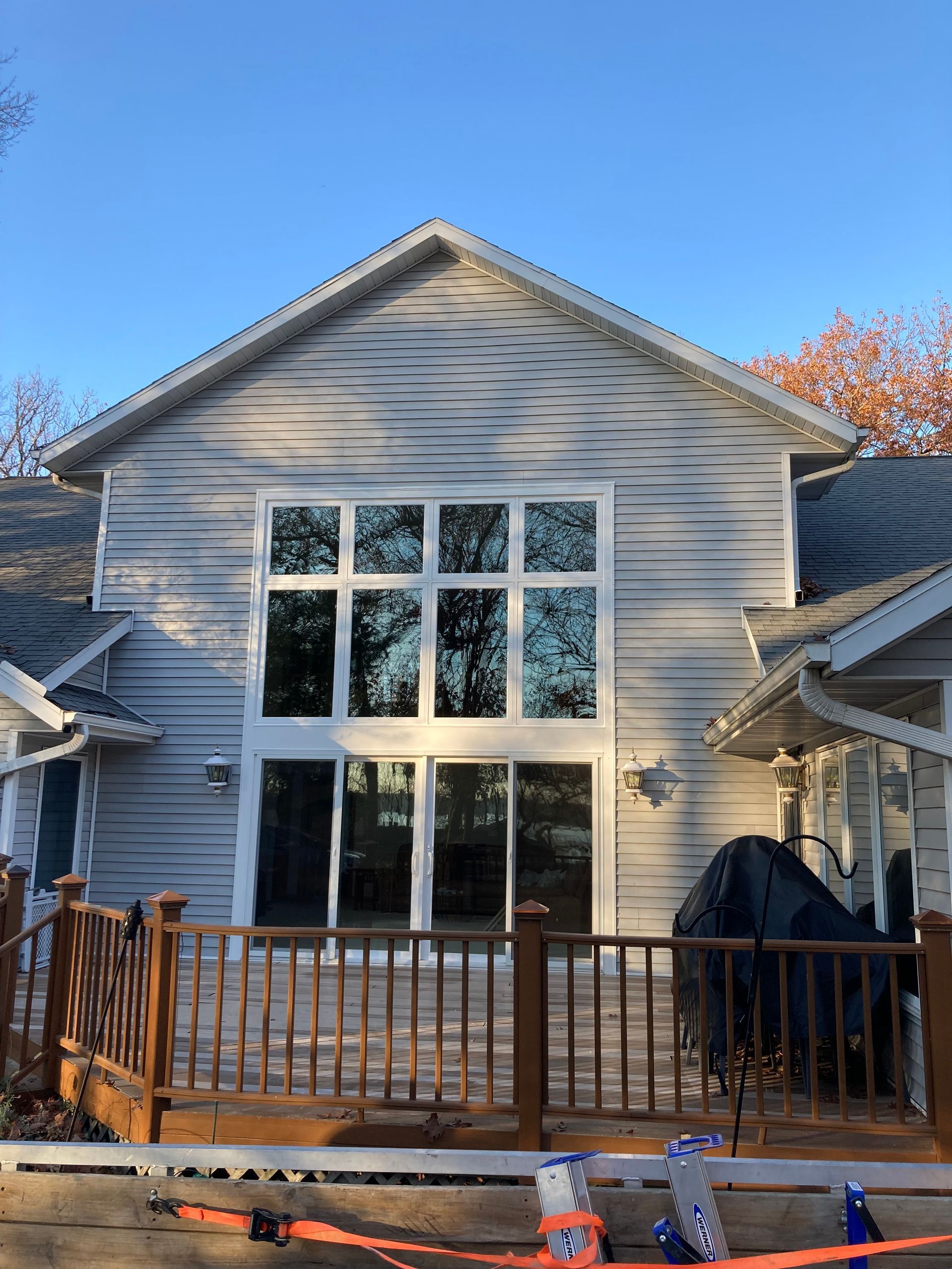 Rear view of a gray house with large windows and a wooden deck, under a clear blue sky.