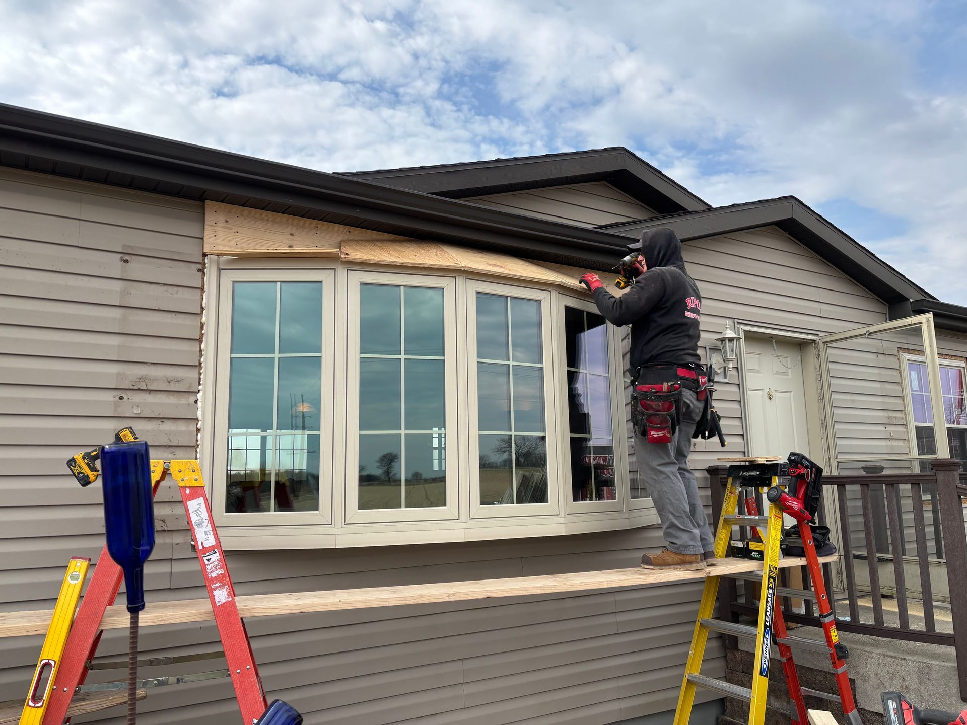 Workers installing a curved window frame on a house porch, with tools and ladders nearby.