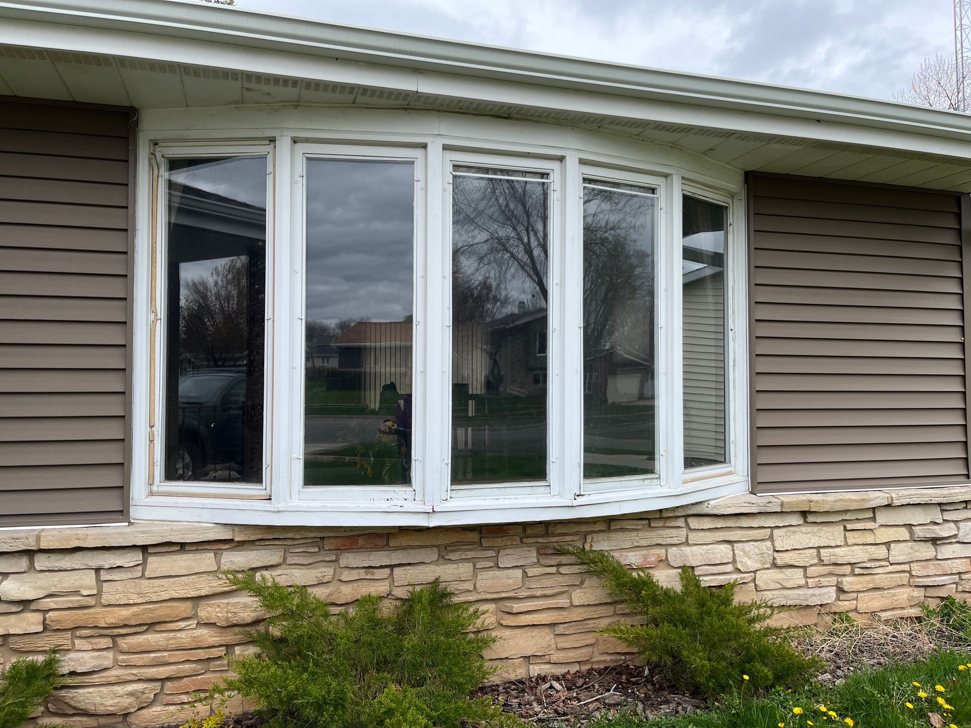 Front of a house with a large white bay window above a stone foundation and beige siding