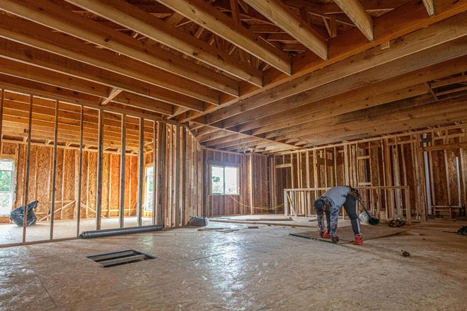Unfinished house interior with exposed wooden framing and a worker bending over near the floor.