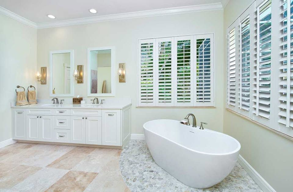 Bright white bathroom with double vanity, mirrors, plantation shutters, and a freestanding soaking tub.