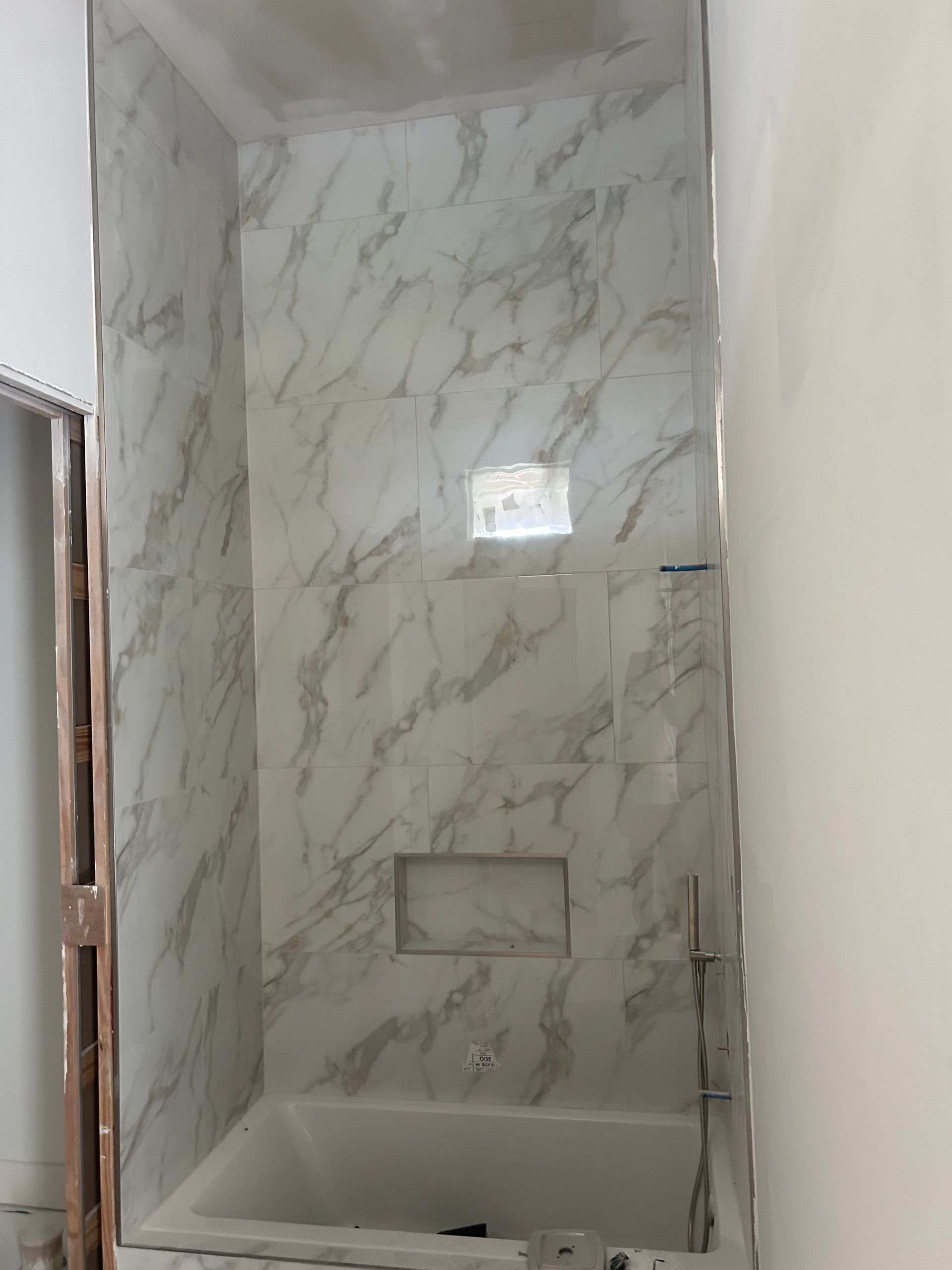 Modern tiled shower stall with white marble-look walls, built-in shelf, and bathtub below