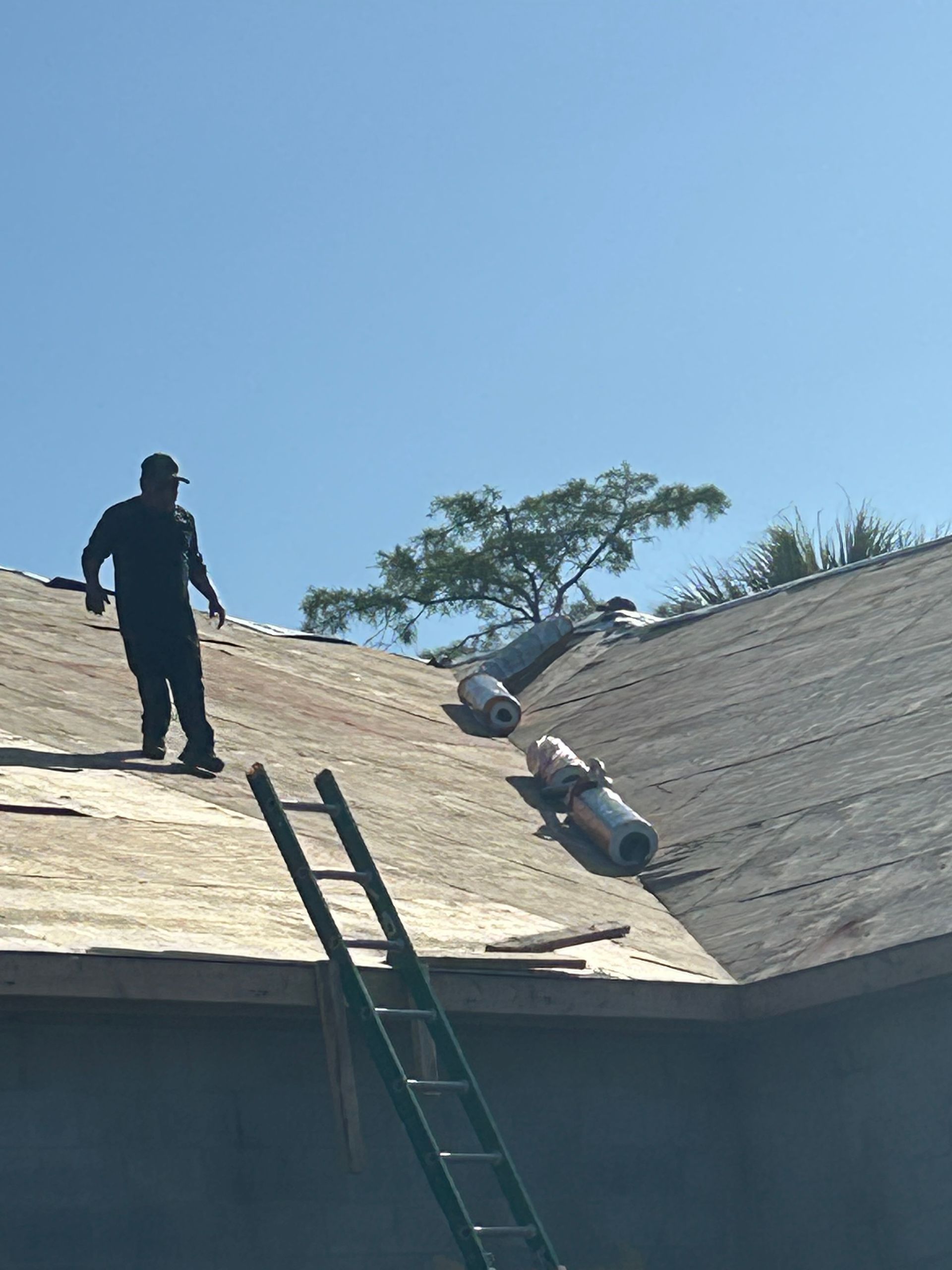 Worker on steep shingled roof beside a ladder and safety bags under a clear blue sky