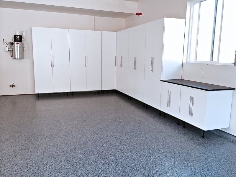 White storage cabinets and a counter in a clean garage with speckled epoxy floor and window light.