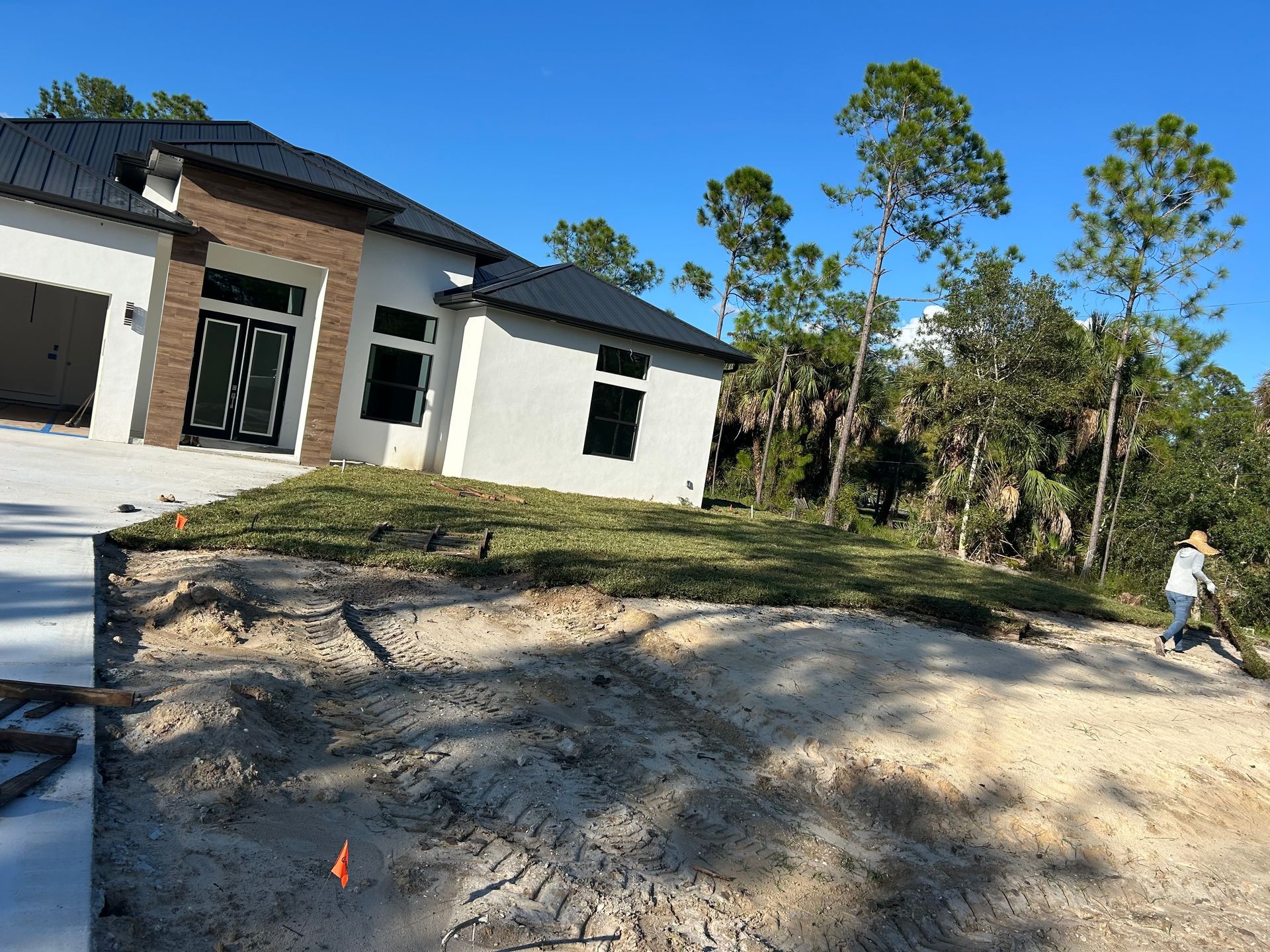 Construction site with a house under renovation, dirt foreground, and a person walking near trees on the right.