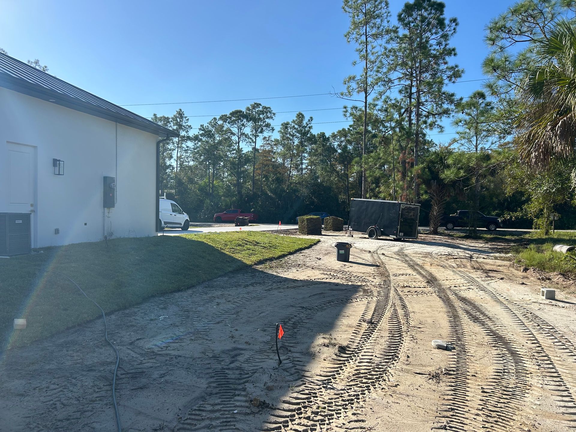 Sandy construction site beside a white building, with tire tracks and trees under a clear blue sky