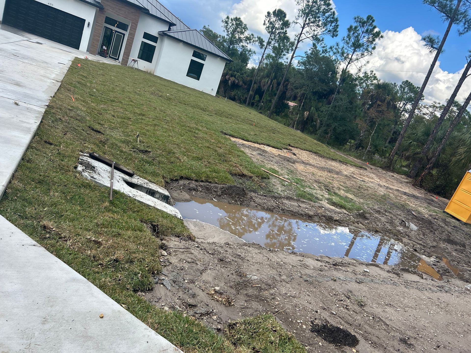 Tilted view of a small white house, lawn, driveway, and muddy puddles after rain
