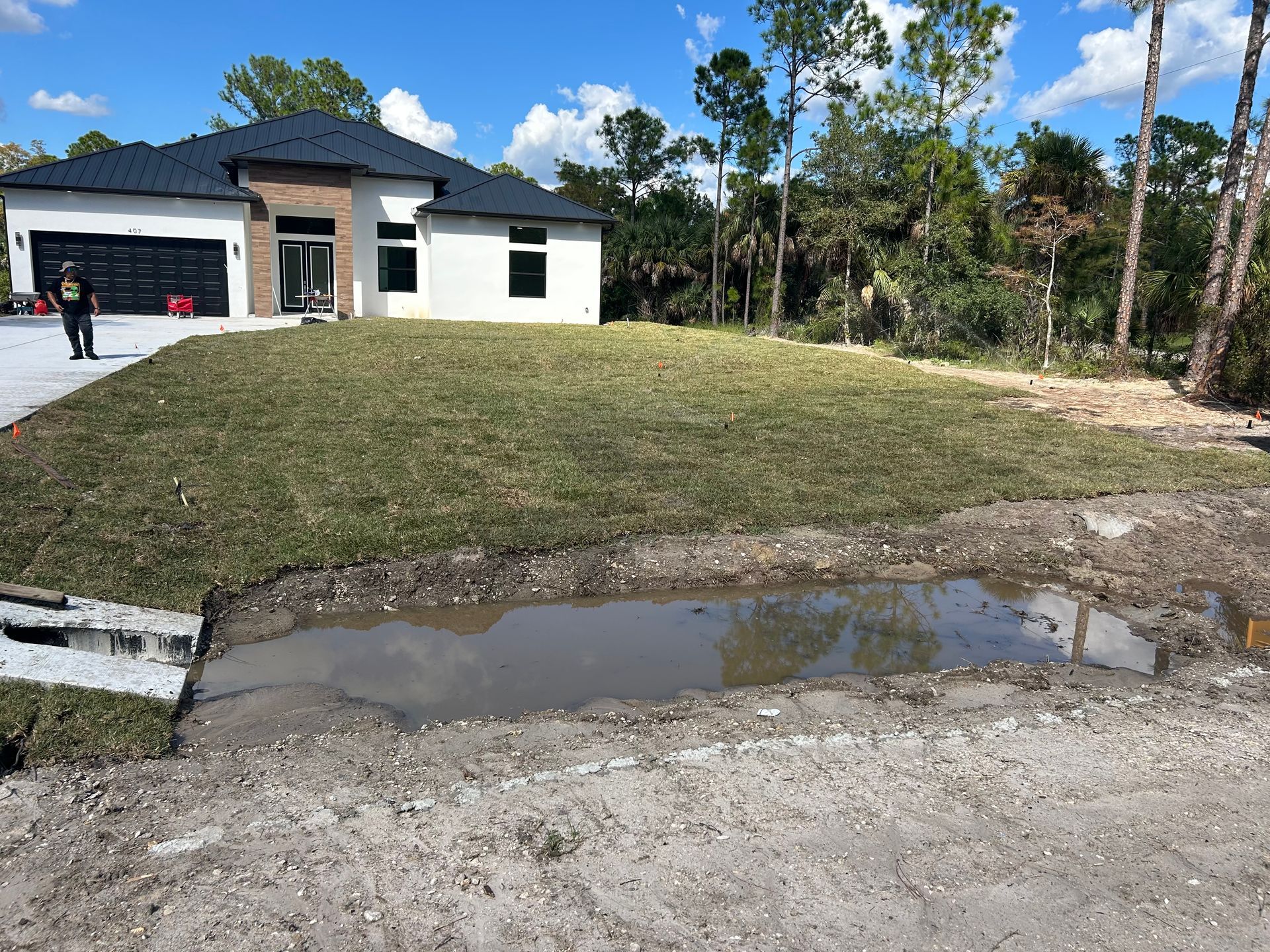 Single-story house with a driveway, large sloped lawn, and a puddle-filled curb under a bright blue sky