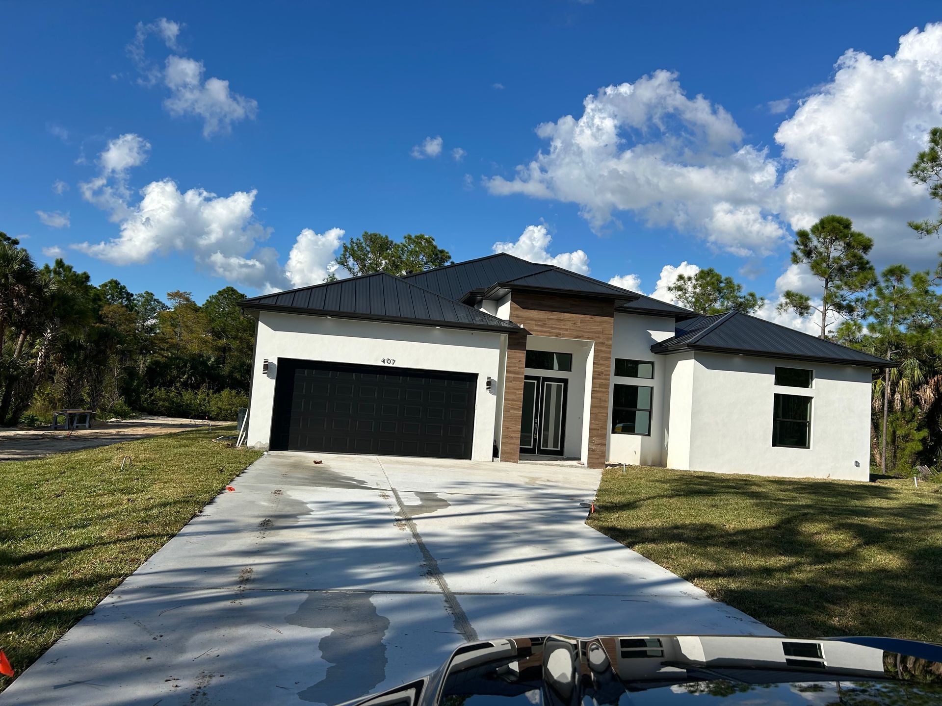 Modern white house with dark roof, black garage doors, and a long concrete driveway under a blue sky.