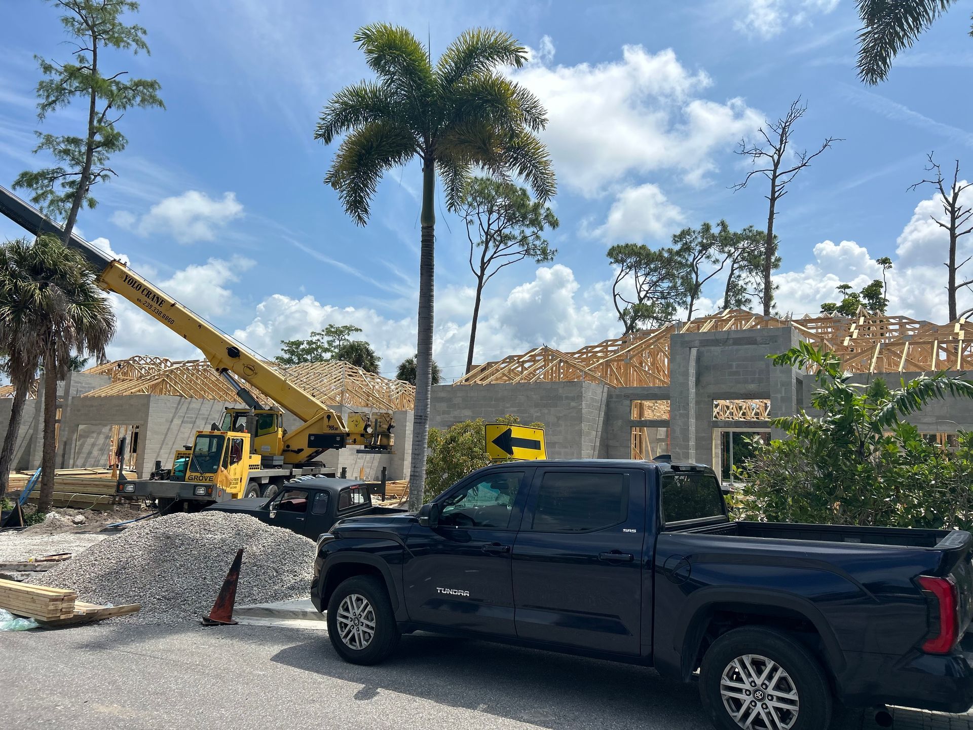 Construction site with yellow crane, black pickup truck, palm trees, and a partially built house under a blue sky