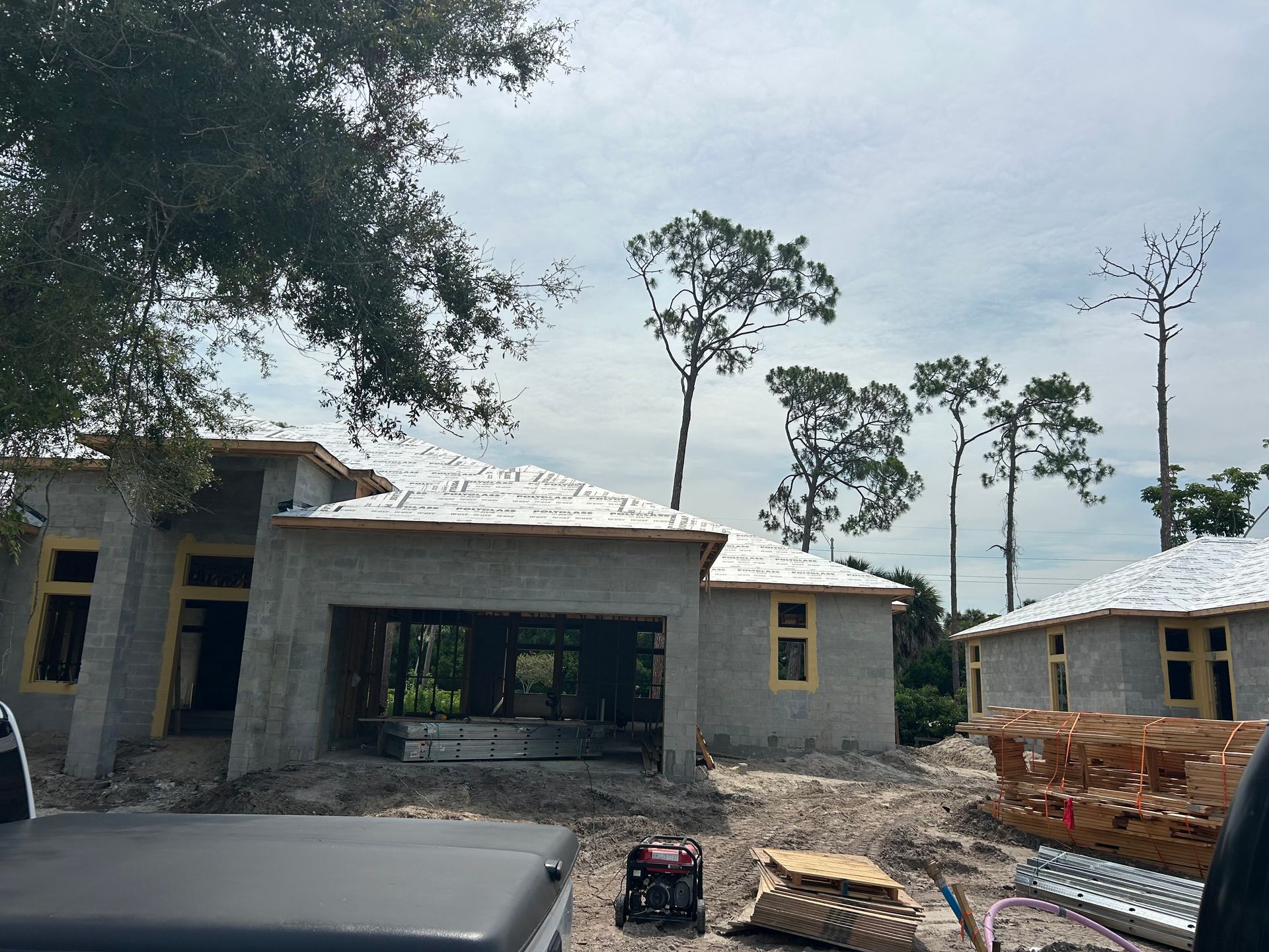 Partly built concrete houses under construction on a dirt lot with stacked bricks and trees nearby