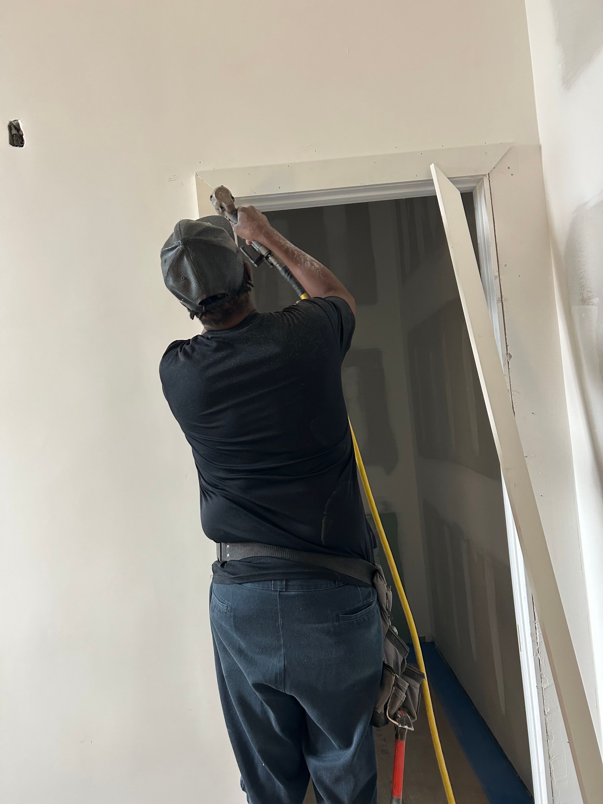 Worker installing trim above a doorway in an unfinished hallway, using a ladder and power nailer.