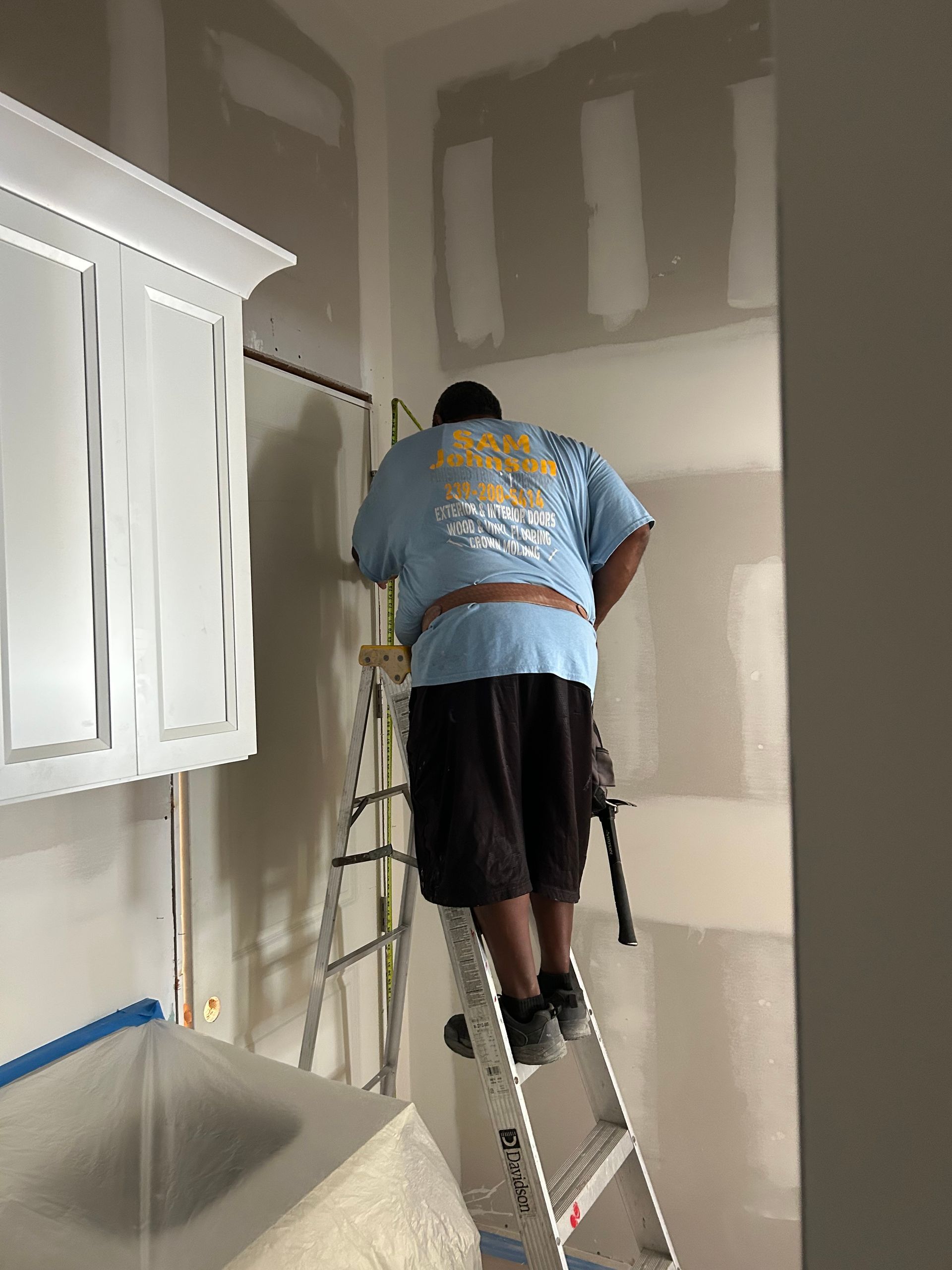 Person standing on a ladder painting unfinished drywall inside a room.