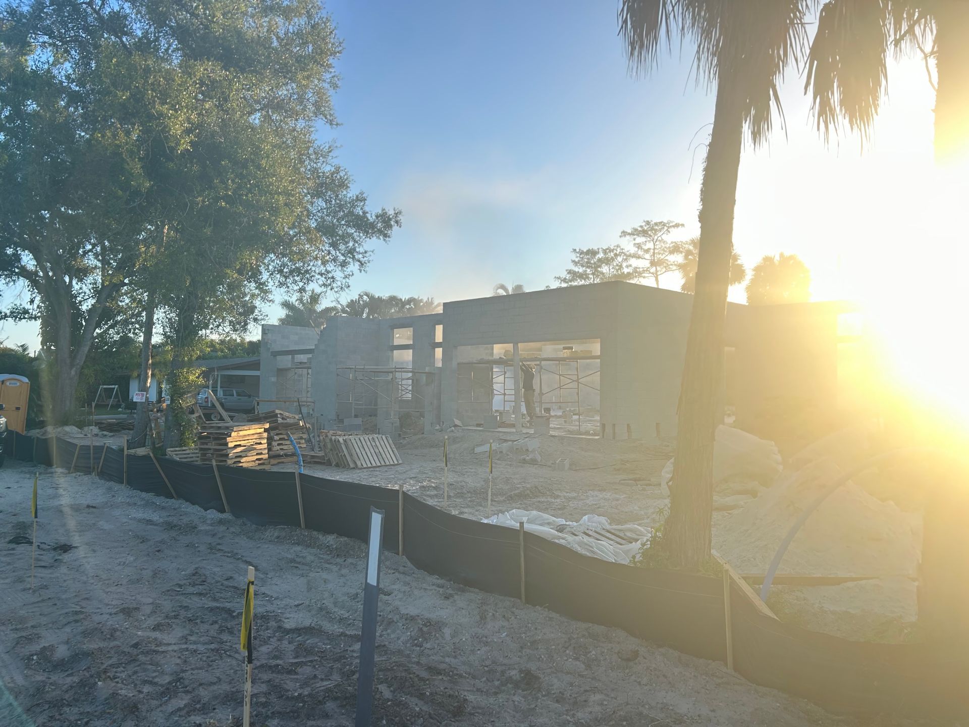 Sunlit construction site with a low cinderblock building, dirt lot, trees, and a safety fence