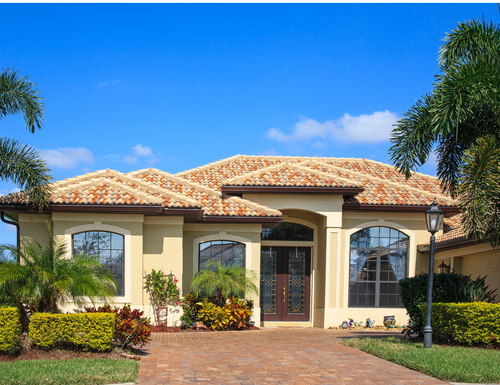 Single-story stucco house with a tiled roof, arched windows, and a brick driveway under a blue sky
