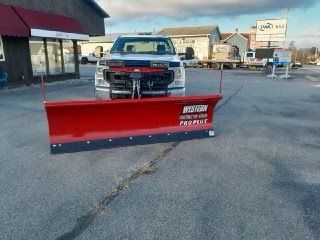 Red snow plow attached to the front of a white truck in a parking lot.