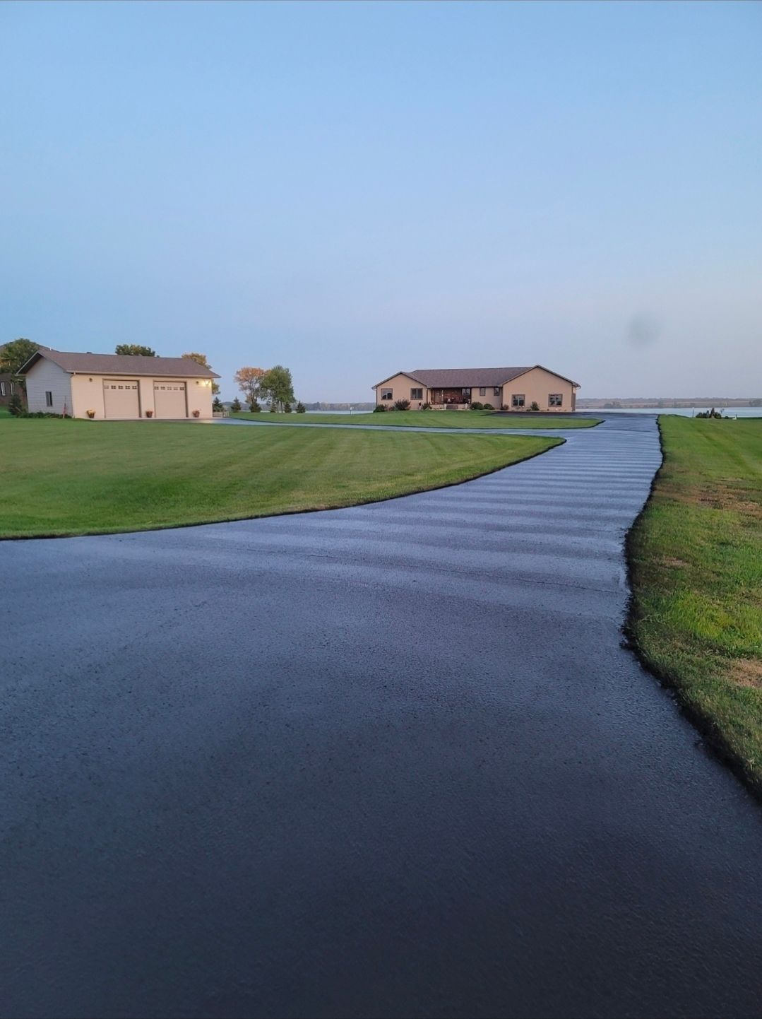 Curving wet driveway through a grassy field toward distant houses under a pale blue sky