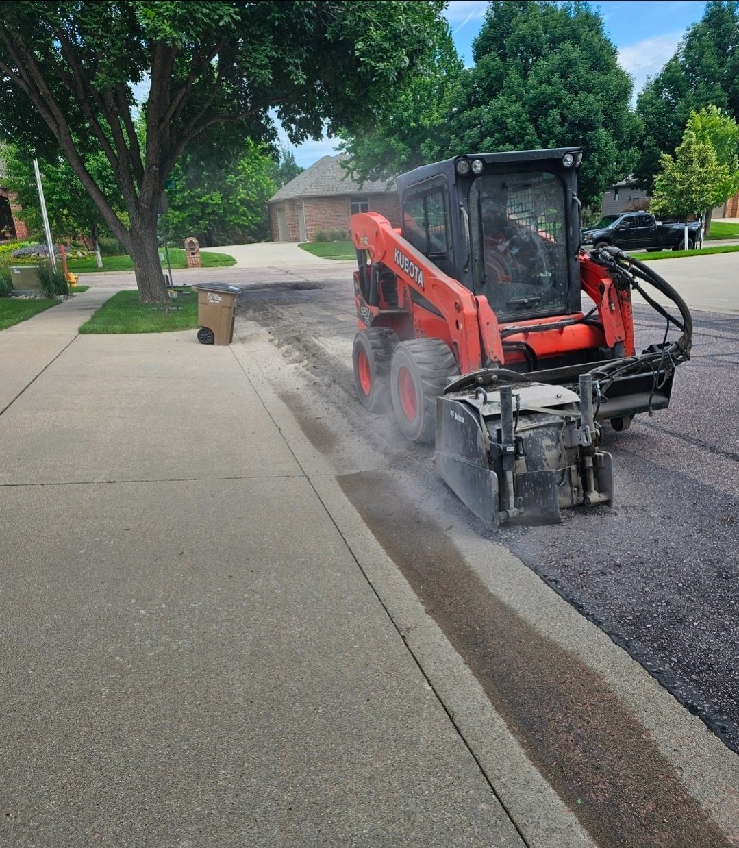 Red skid-steer sweeping dust from a curb beside a sidewalk on a suburban street