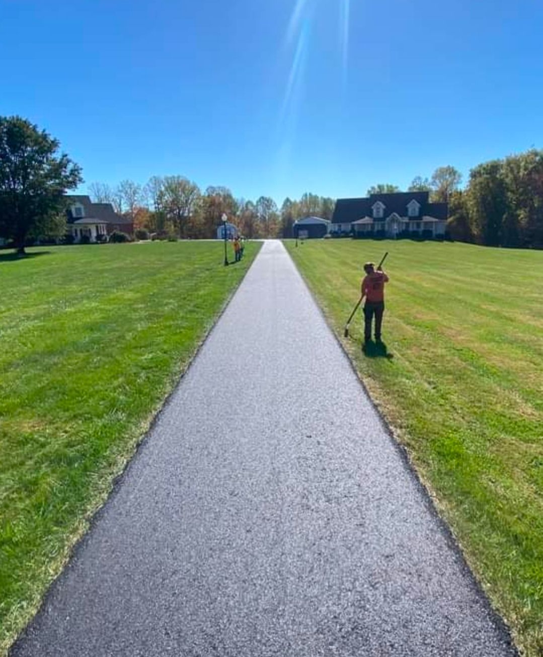 A long paved path across a green lawn with two people standing on either side under a blue sky.