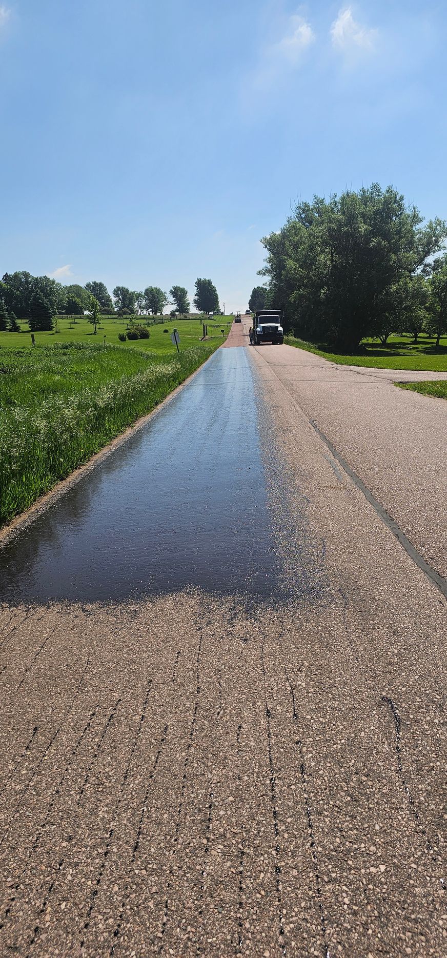 Wet rural road beside green fields under a bright blue sky, with a person walking in the distance
