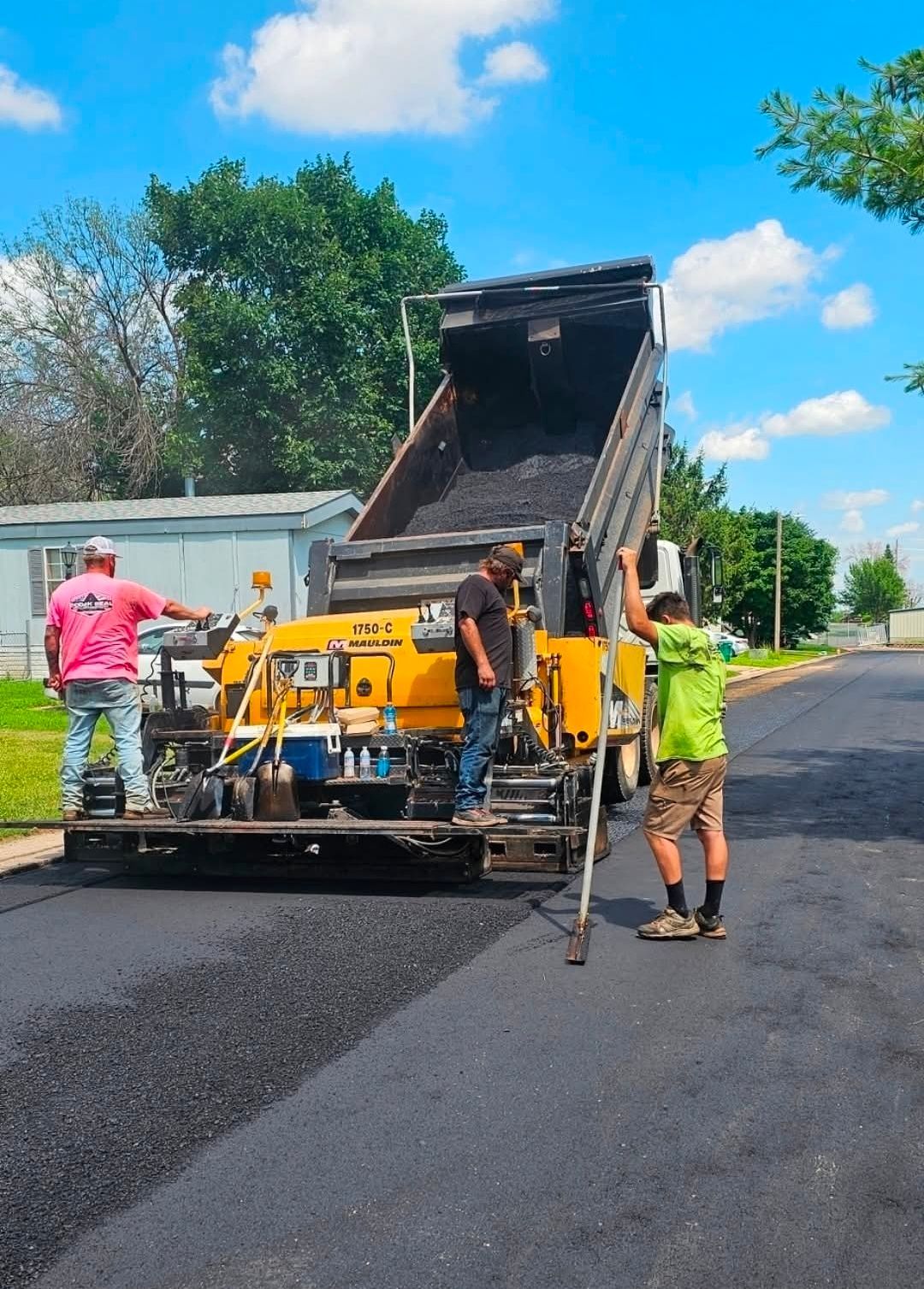 Workers paving a road as a dump truck empties asphalt onto a road paver under a blue sky