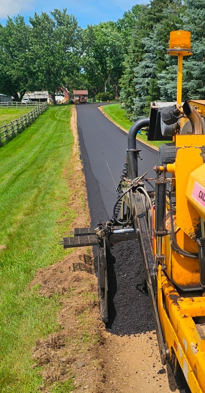 Yellow paving machine laying black asphalt along a grassy roadside path