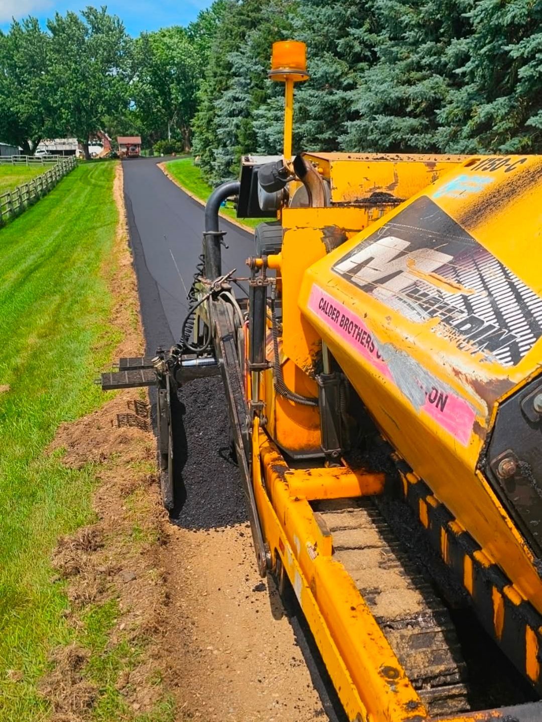Orange farm machine working beside a paved path and green field on a sunny day