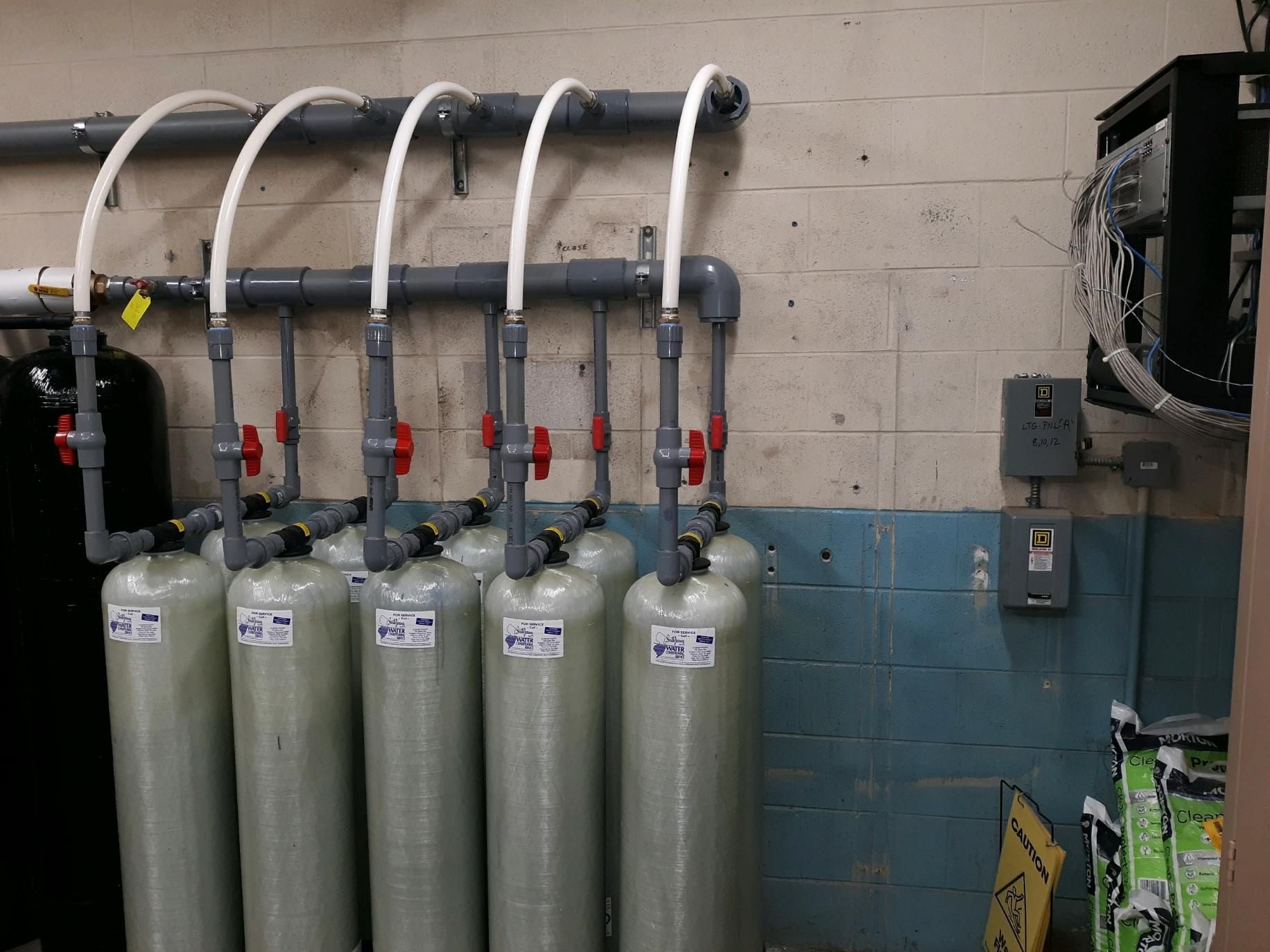Row of gray industrial gas cylinders with pipes and valves against a blue and beige wall