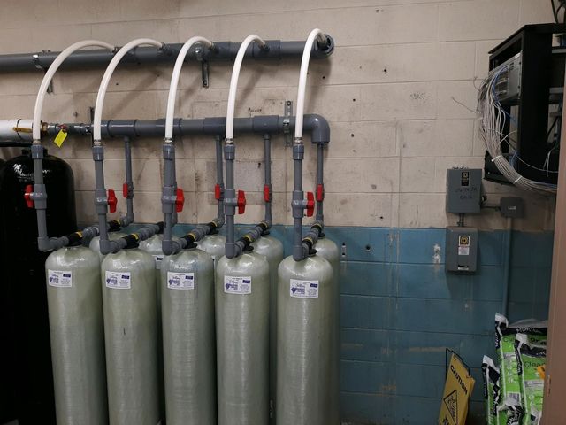 Five upright gas cylinders with hoses and valves in an industrial room, next to a wall-mounted control panel.