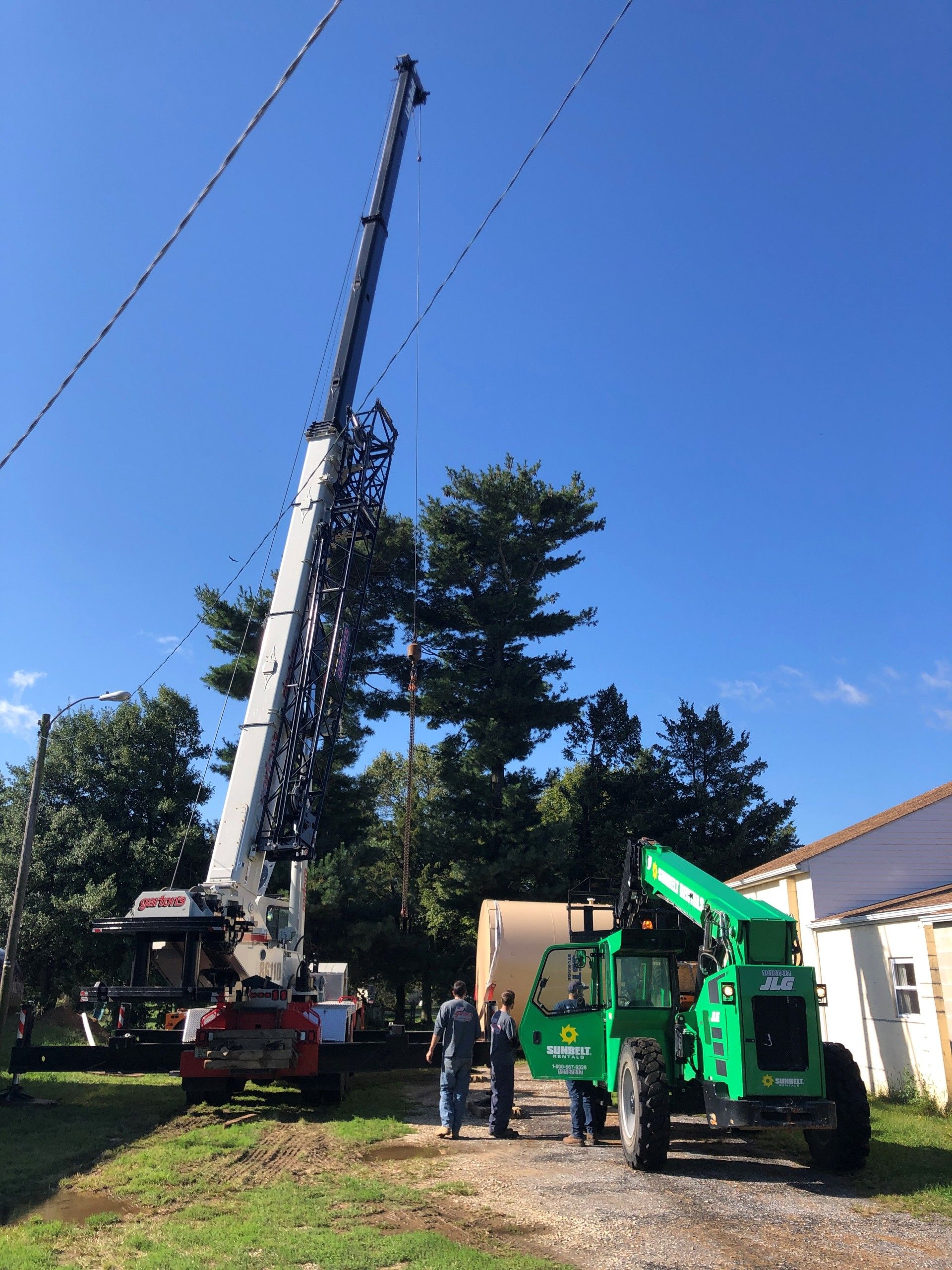 Crane lifting a tall tree beside a house, with workers and a green forklift in the yard.