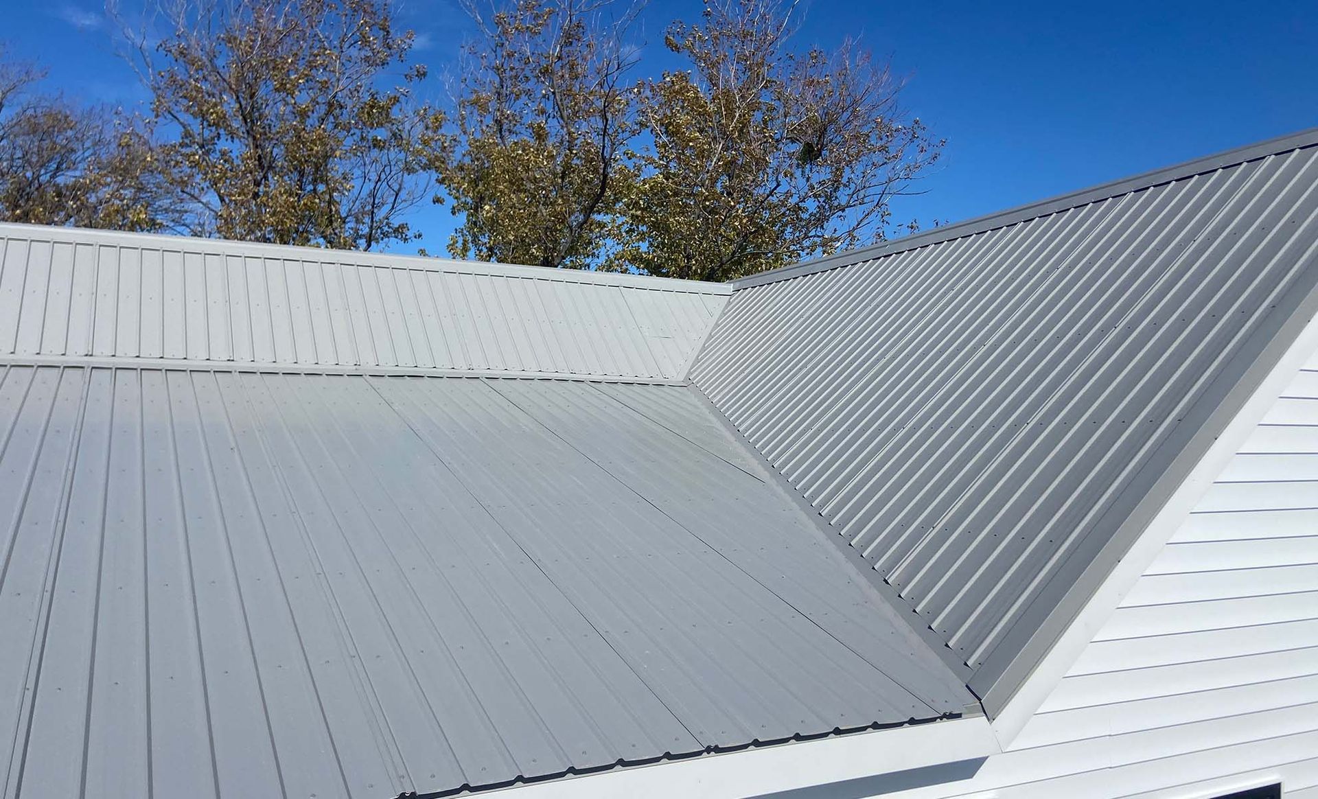 White metal roof with a steep seam and blue sky above trees