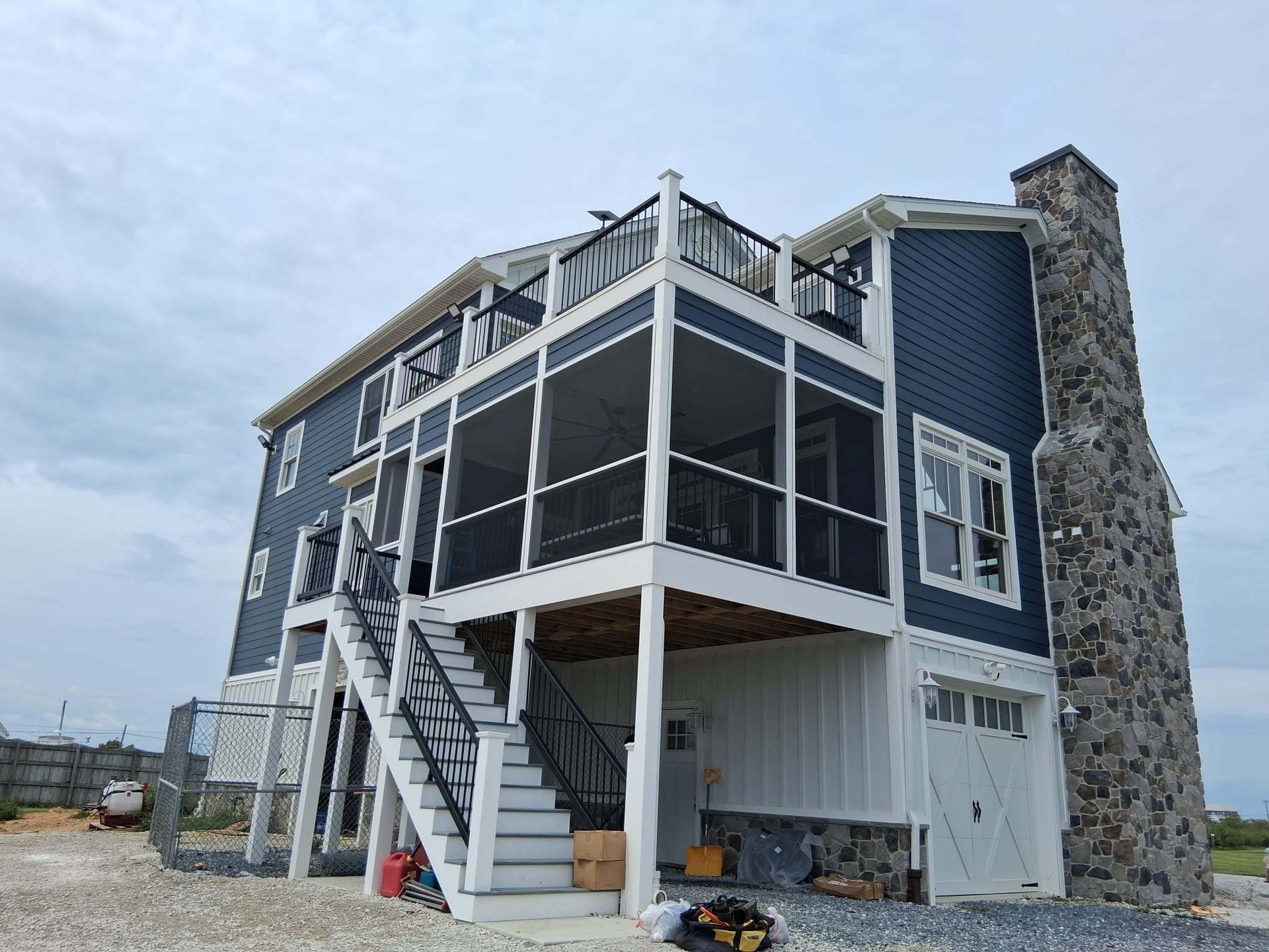 Blue coastal house with screened porch, white stairs, and stone chimney under cloudy sky