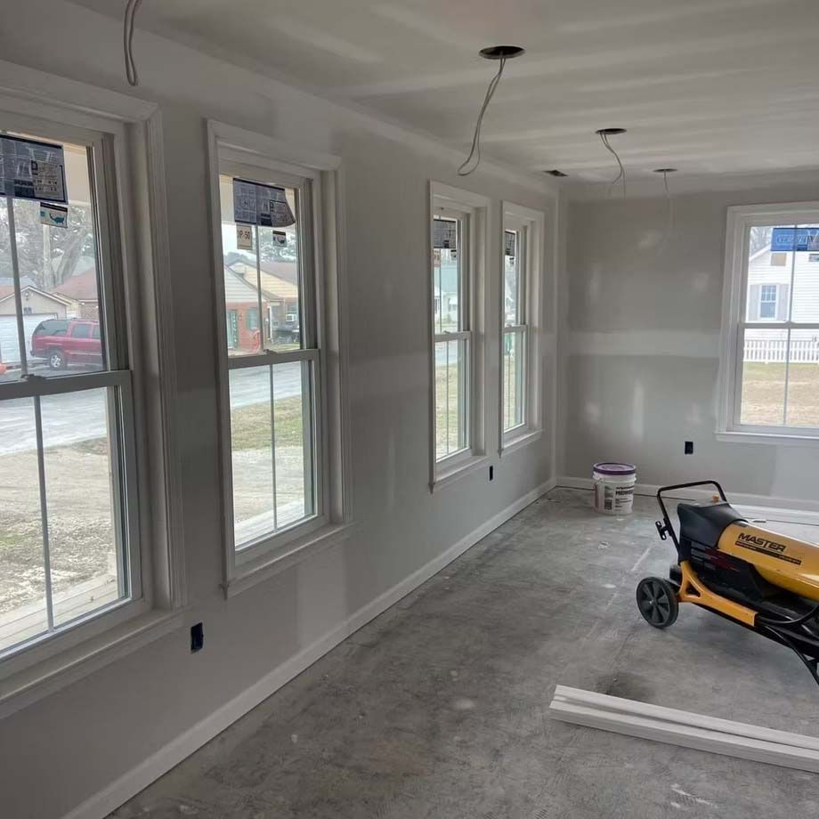 Bright sunroom under construction with white walls, multiple windows, unfinished floor, and a yellow lawn mower inside.