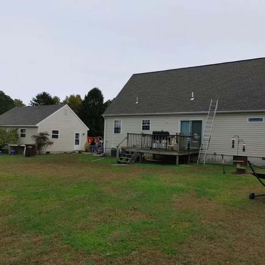 Suburban backyard with a beige house, wooden deck, ladder, and a small shed under an overcast sky