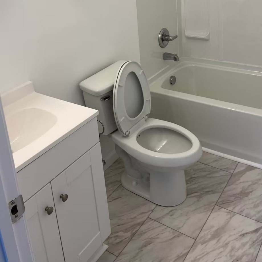 Small bathroom with toilet, sink vanity, bathtub, and gray tile floor; toilet seat is open.