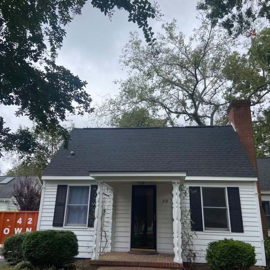 Small white house with dark roof, black shutters, front porch, and brick chimney under trees
