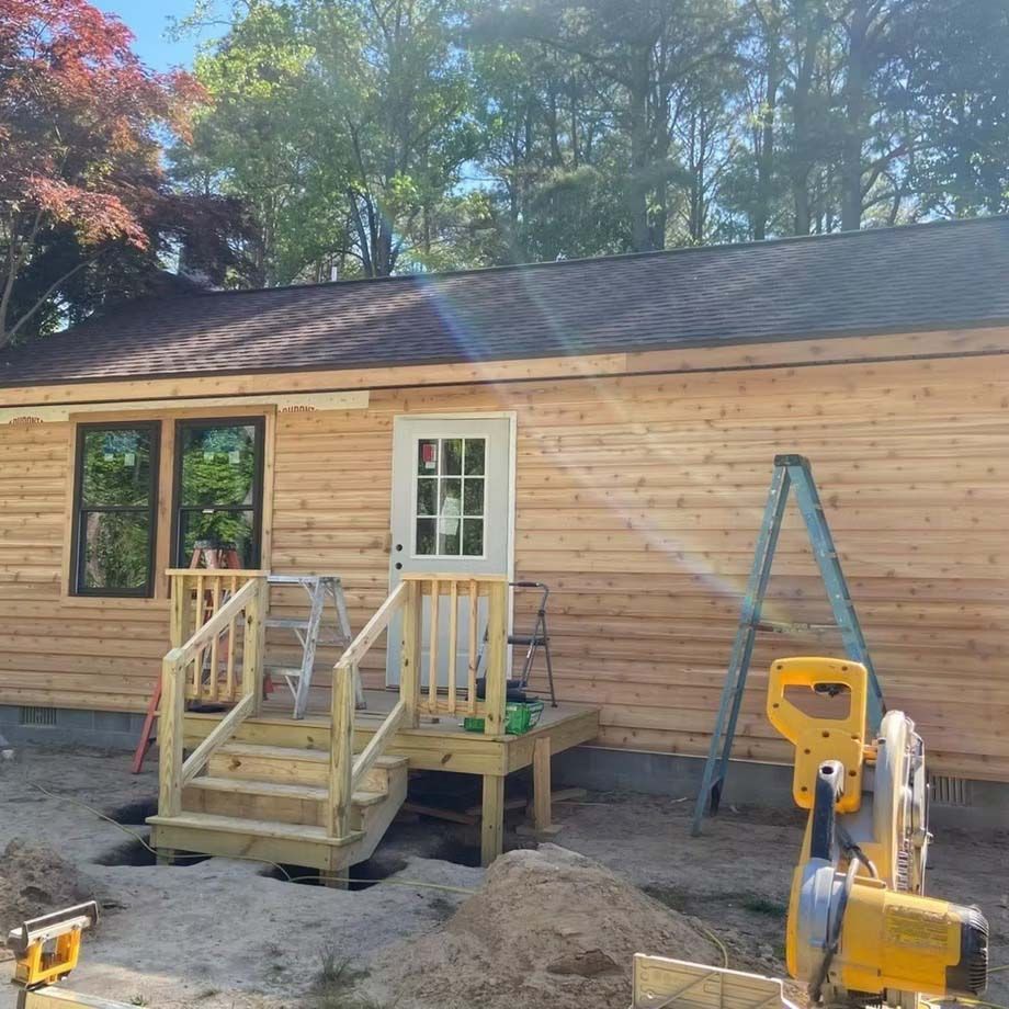 Wood-sided house exterior with small front deck, stairs, ladder, and construction tools in the yard
