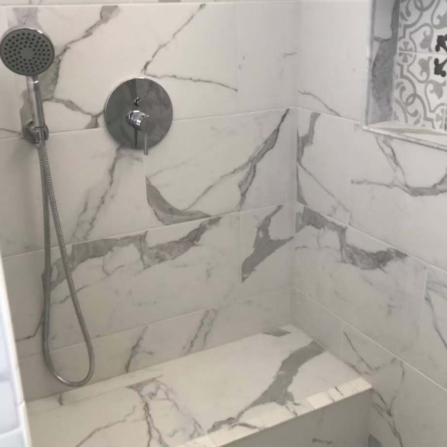 Modern shower with marble-look tile, wall-mounted controls, handheld showerhead, and built-in bench