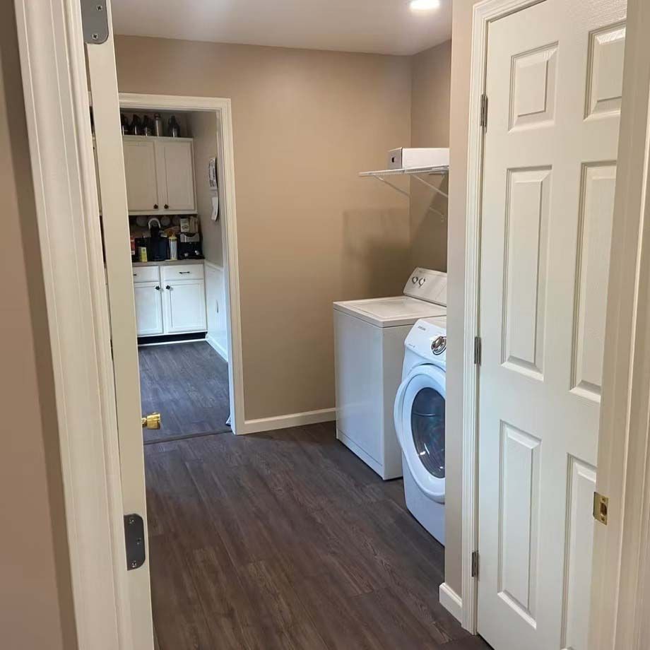 Narrow laundry room with washer, shelves, and an open doorway to a kitchen beyond
