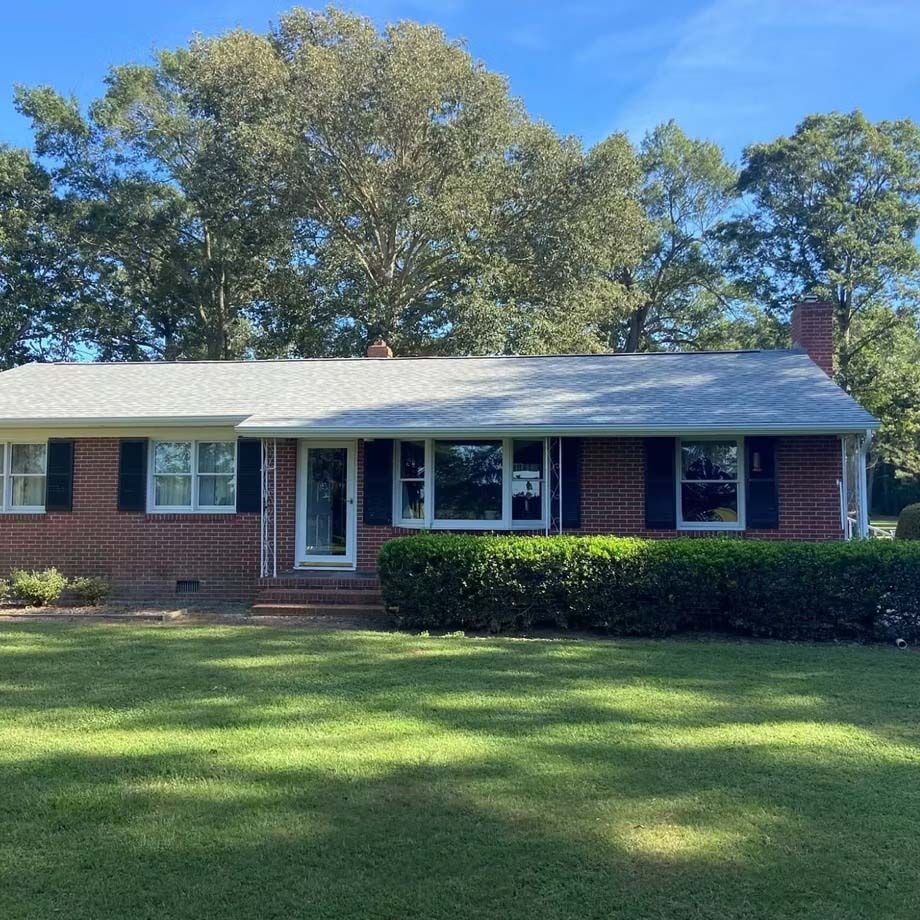 Single-story brick house with white trim, front porch, and large green lawn under a blue sky